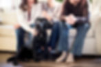 Three people sit on a beige couch with a black Labrador dog. The scene is warm and cozy with natural light, creating a joyful mood.