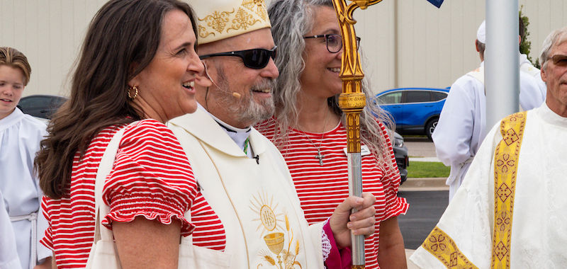 A bishop, wearing white with ornate gold details, stands smiling with two women in red-striped shirts outdoors, near a flag and other clergy.