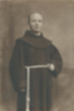 Sepia portrait of a smiling monk in a brown robe with a rope belt, holding a rosary against a plain studio backdrop