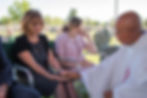A somber outdoor gathering at a cemetery. A woman in a black polka-dot dress holds a priest's hand. Another woman in pink looks on.
