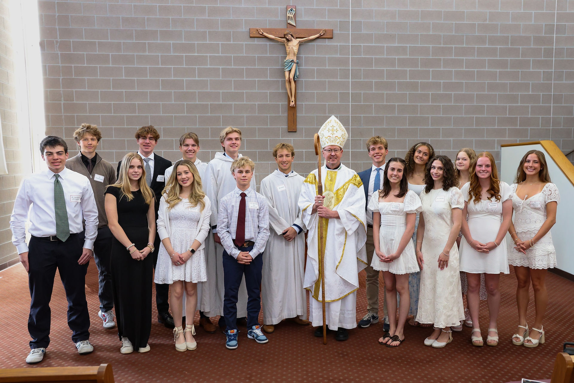 Catholic bishop poses with high school students beneath a large crucifix following a school Mass celebration.