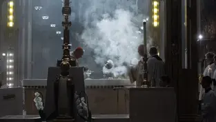 Clergy perform a ritual with incense smoke in a dimly lit cathedral. Ornate altar with white flowers; solemn and reverent atmosphere.