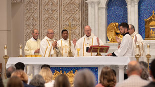 Clergy in white vestments celebrate Mass at a gold altar before a blue mosaic backdrop, with worshippers seated in front.