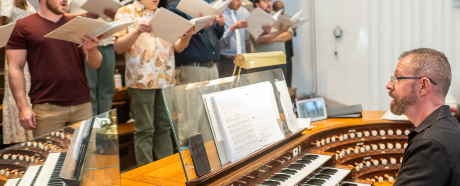 Choir singing with sheet music as an organ player performs. Wood-paneled background, warm colors, and a focused, harmonious mood.