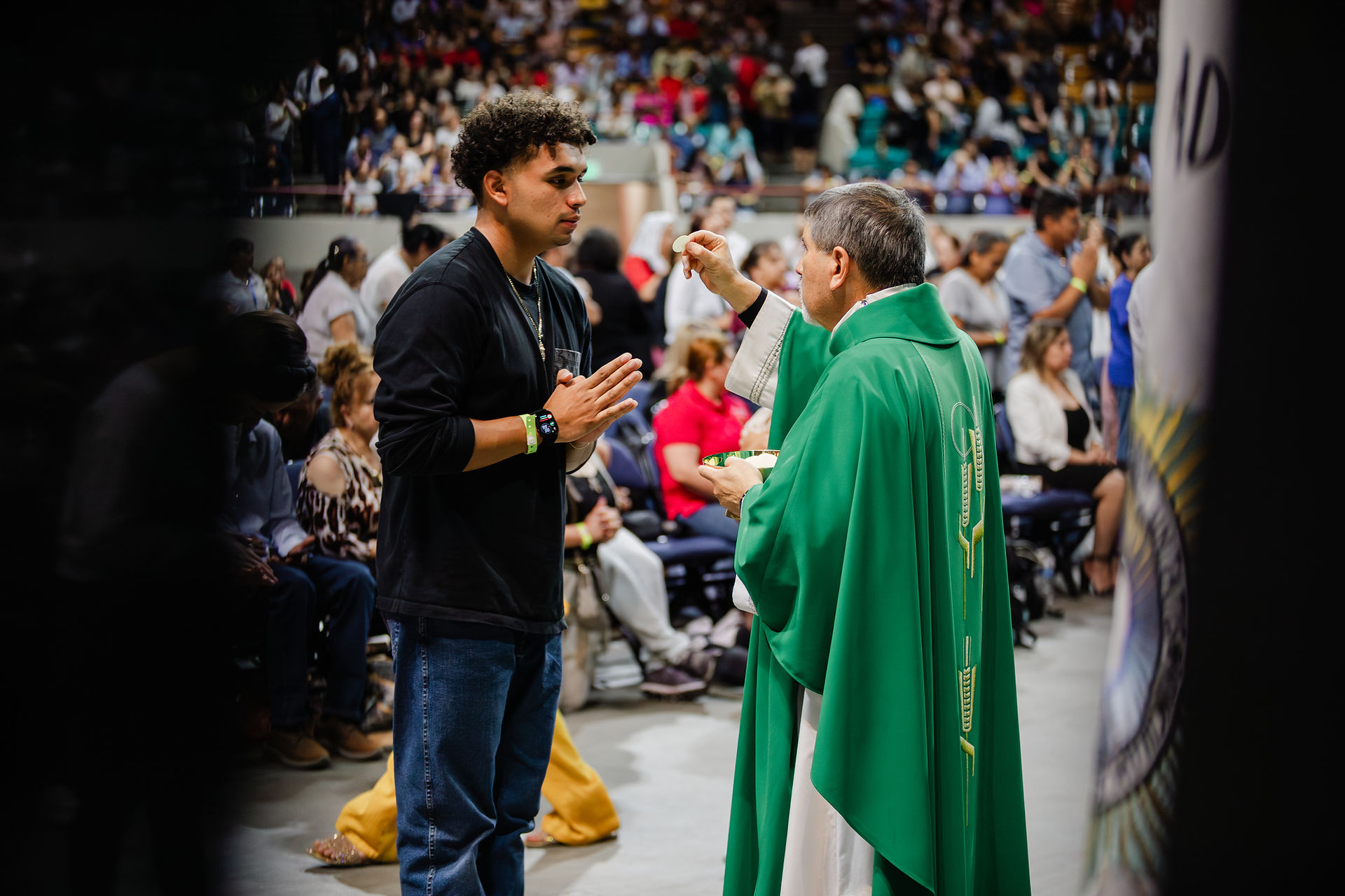 Priest in green robe holds Communion wafer before a man with clasped hands. Crowd in background; solemn church setting.