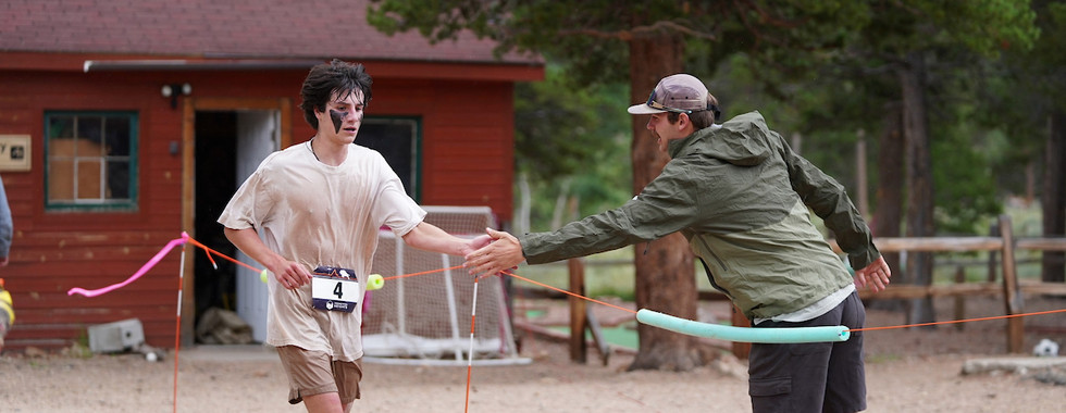 Runner with face paint in dirty clothes approaches finish line, high-fiving a man in a cap. Red cabin and trees in the background.