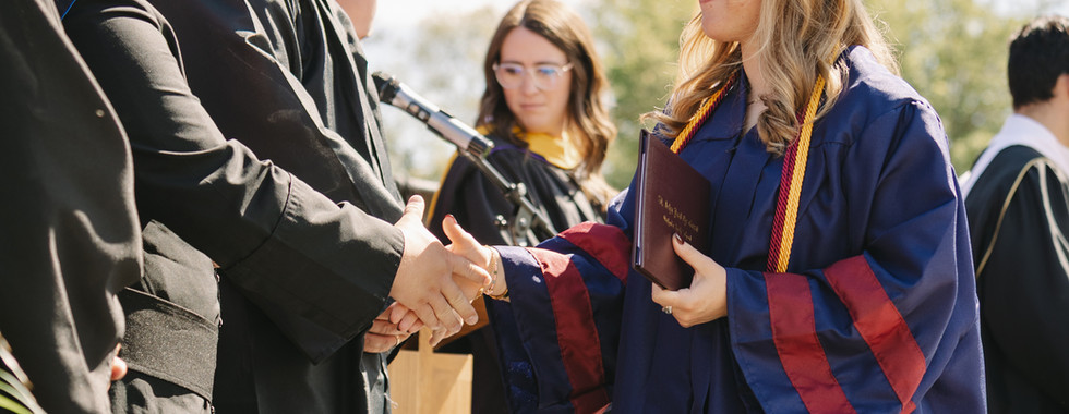 Graduate in blue cap and gown shakes hands with a faculty member at an outdoor commencement ceremony, holding a diploma folder.