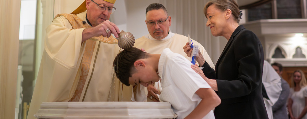 A bishop pours water over a child’s head during a baptism while a woman stands beside them smiling.
