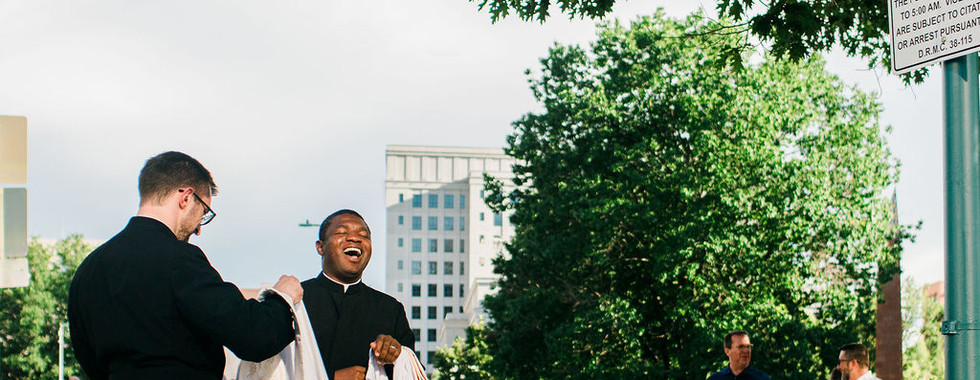 Two priests in black robes laugh on a tree-lined park path, with people walking by and a posted closed-area sign on a green lamppost.