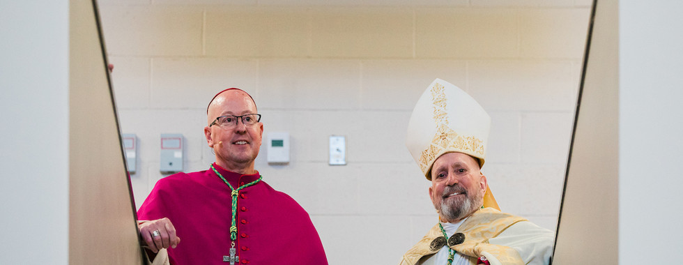 Two clergymen in ceremonial robes and hats stand on stairs, smiling. The wall behind them is beige with visible electrical panels.
