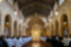 Clergy in white robes process in a grand church with arched ceilings and stained glass. A solemn ceremony unfolds at the ornate altar.
