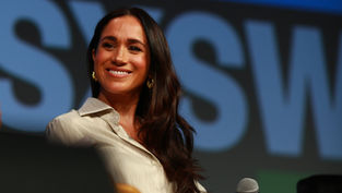 Smiling woman with long brown hair wearing a light shirt, on stage with "SXSW" text in background. Warm lighting, microphone visible.
