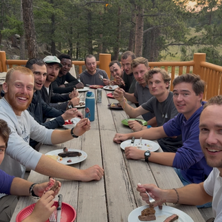 Group of people enjoying a meal on a wooden deck, surrounded by trees. Everyone is smiling and holding utensils, creating a cheerful mood.