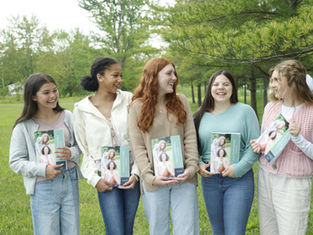 Five smiling women holding books titled "Friendships" stand outdoors on grass with trees in the background, all wearing casual outfits.