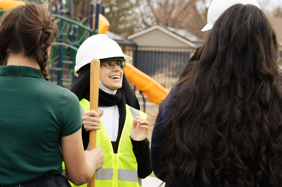 Smiling nun in hard hat and neon vest talks with two women at a playground, holding a shovel near an orange slide.