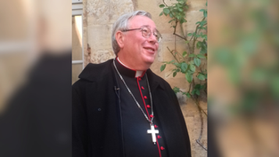 A smiling man in clerical attire with a cross necklace stands outdoors, beige stone wall and greenery in the background.