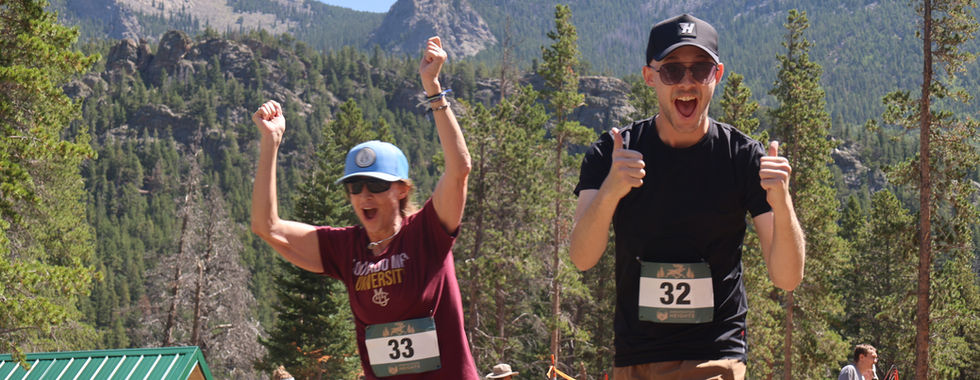 Two runners with bibs 33 and 32 cheer and give thumbs-up on a mountain trail, with pine trees and rocky peaks behind them.