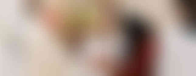 A bishop in ornate robes receives a bowl from a woman in a veil. Altar servers hold candles in a church setting with soft lighting.