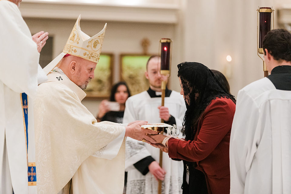 A bishop in ornate robes receives a bowl from a woman in a veil. Altar servers hold candles in a church setting with soft lighting.