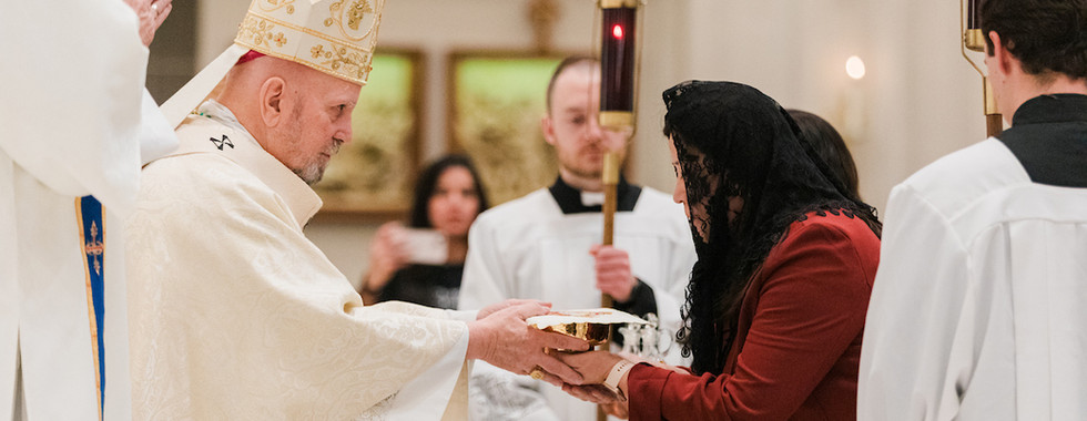 A bishop in ornate robes receives a bowl from a woman in a veil. Altar servers hold candles in a church setting with soft lighting.