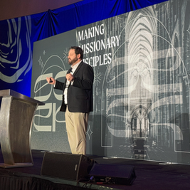 Man speaking on stage with a microphone, in front of a backdrop reading "Making Missionary Disciples." Blue lighting and cathedral image.