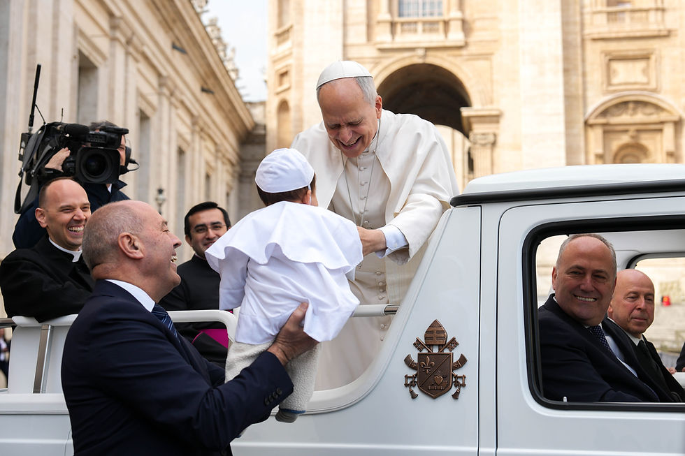 A smiling religious leader in white greets a child in a white outfit, held by a smiling man. They're near an ornate building, filmed by a cameraman.