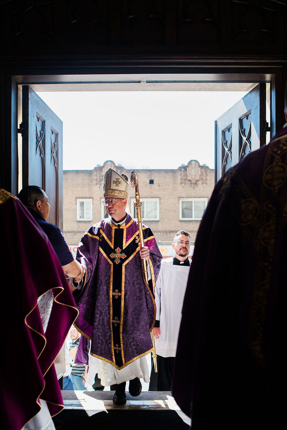Obispo entra por las puertas de la iglesia mientras saluda a una persona, acompañado por ministros.