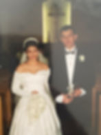 Bride and groom in a church, standing side by side. The bride in a white dress with flowers, the groom in a black tuxedo. Cross visible behind.