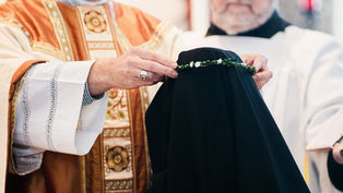 A person in ornate robes places a green floral crown on a figure in black cloth. The setting is solemn and ceremonial.