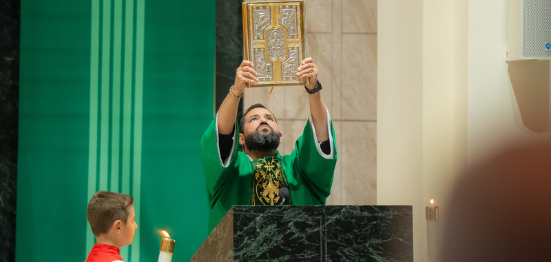 Deacon in green vestments lifts a gold book at the altar while an altar boy stands beside a candle in a solemn church interior.