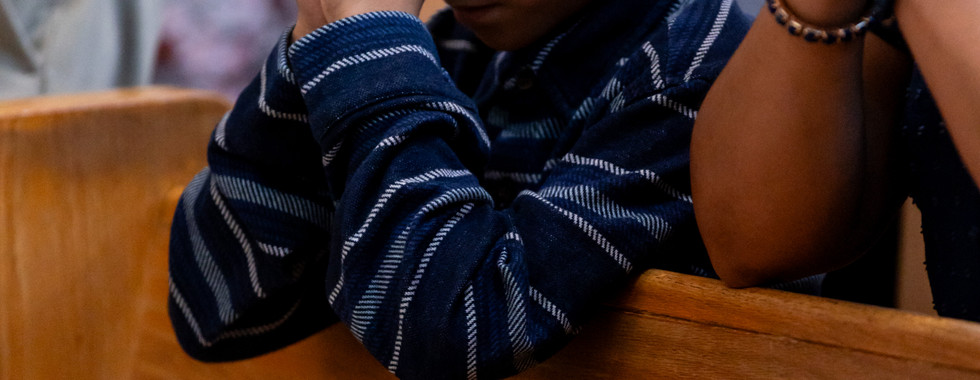 Boy in striped shirt prays with clasped hands in a wooden church pew, surrounded by others with similar gestures. Serene atmosphere.