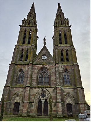 Twin-spired Gothic cathedral facade with pointed arches and clock, against a cloudy sky. Weathered stone and green moss detail.