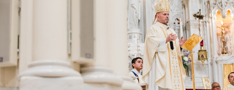 Bishop in ornate white and gold robes speaks at a lectern in an ornate church setting with carvings and religious figures in the background.