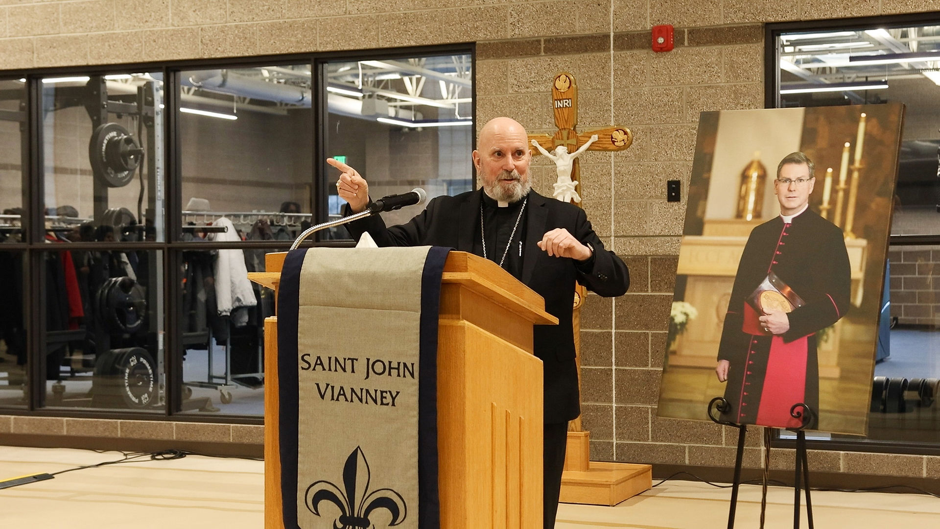 A bishop speaks at a podium labeled "Saint John Vianney" beside a bishop's portrait. Gym equipment and a crucifix are in the background.