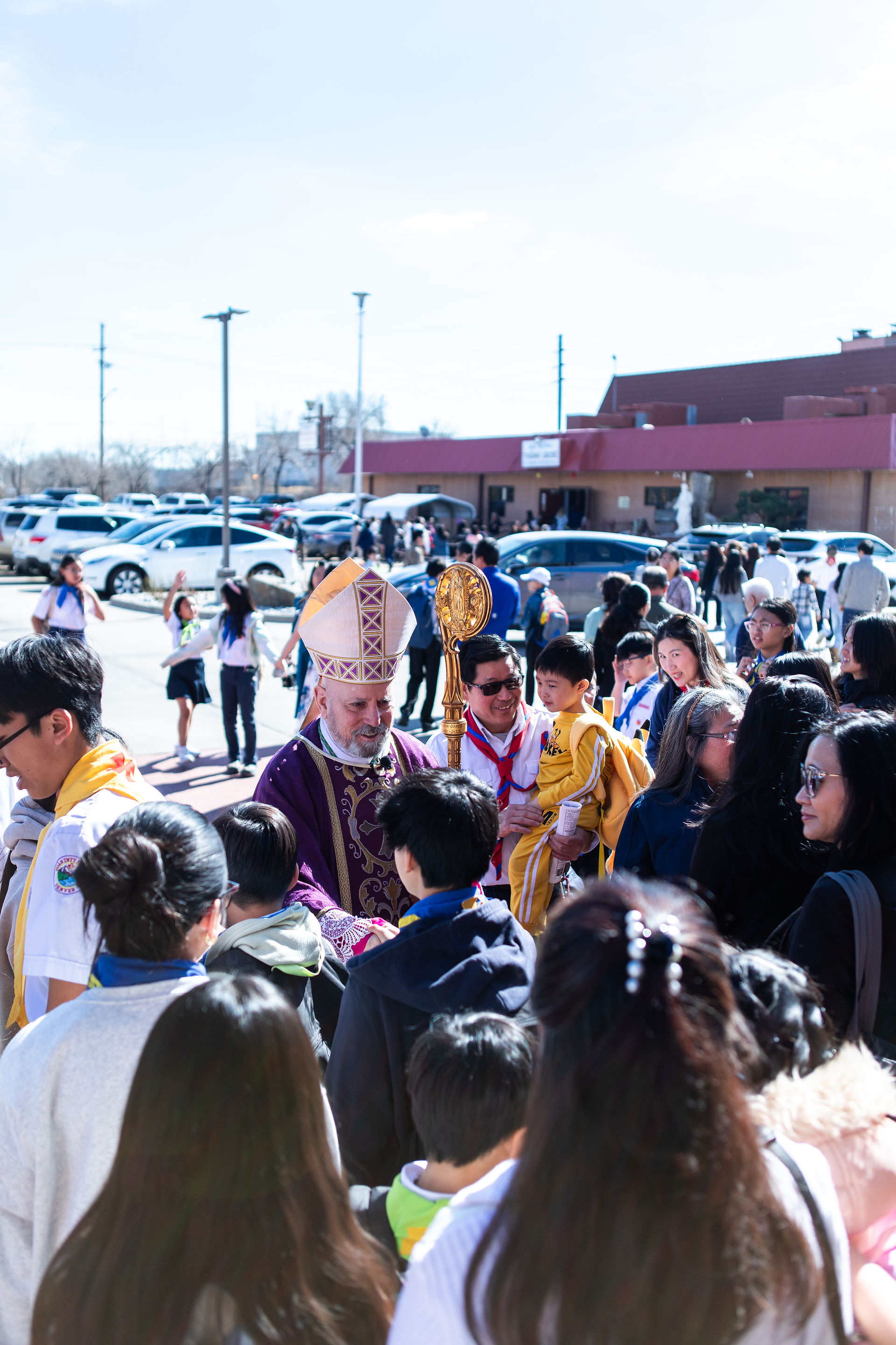 A bishop in purple robes holding a staff greets a crowd outdoors. A man holds a child in yellow. Cars and a building in the background.