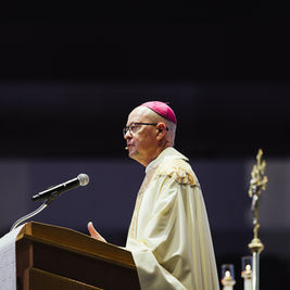 A bishop in ornate robes speaks at a podium with a microphone. He wears a pink zucchetto. Dark background with candle and staff visible.