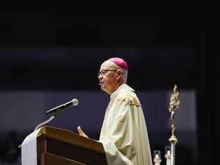 A bishop in ornate robes speaks at a podium with a microphone. He wears a pink zucchetto. Dark background with candle and staff visible.