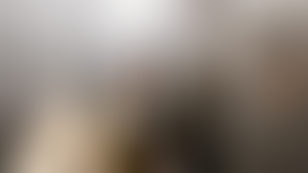 Man in ornate religious attire holds a staff with a cross, under bright light in a dim setting, gesturing with his hand.