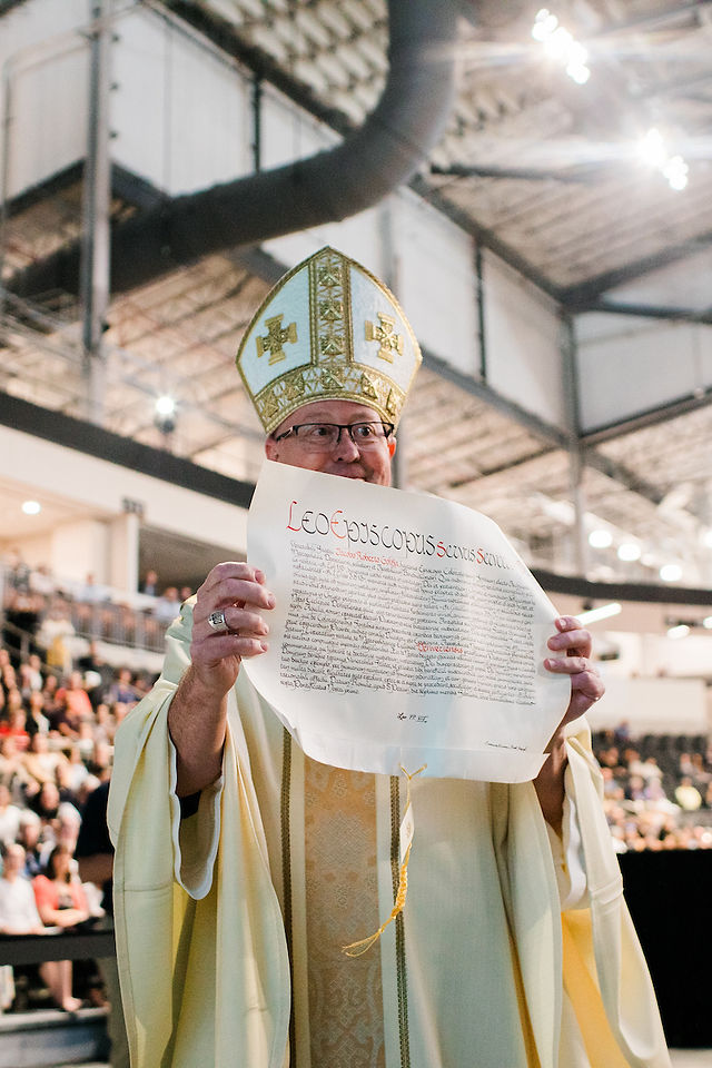 A newly installed bishop holds up an apostolic mandate as the congregation looks on.