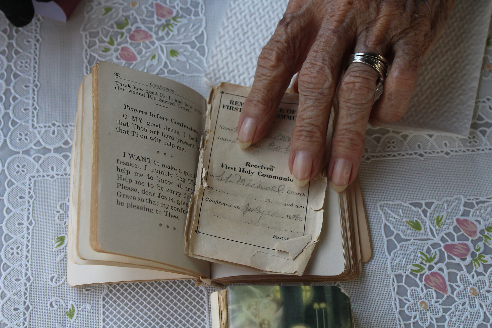 A hand with rings points to a tattered prayer book on a lace tablecloth. Text includes "Prayers before Confession" and a communion record.