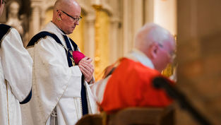 Clerics in ornate robes, one in red, another holding a pink hat, in a church setting with arched decor, conveying a solemn mood.