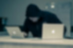 Hooded person working on two laptops at a desk, focused intently. Background shows a whiteboard. Notepad and pen on the desk.