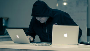 Hooded person working on two laptops at a desk, focused intently. Background shows a whiteboard. Notepad and pen on the desk.