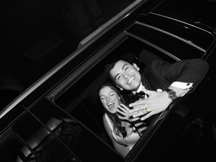 A smiling couple leans out of a car window at night, showing off rings. The man wears a tuxedo. Black and white image. Joyful mood.