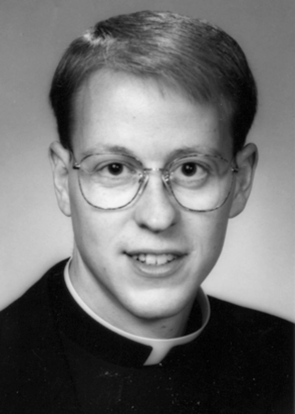 Young person in clerical attire, wearing glasses, smiles at the camera. Black and white portrait with a neutral background.