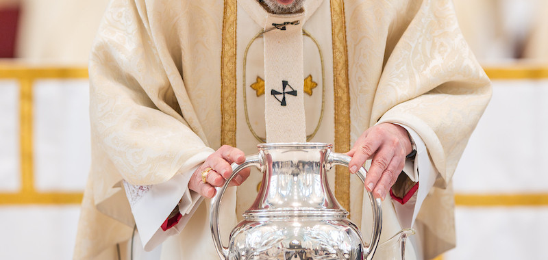 A bishop in ornate robes and a red cap bows over a large silver jug on a wooden table in a church setting, performing a ceremonial act.