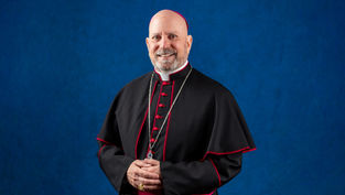 Cleric in black and red robes smiles, standing against a blue background. He wears a gold ring and chain, conveying a serene and formal mood.