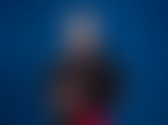 Cleric in black and red robes smiles, standing against a blue background. He wears a gold ring and chain, conveying a serene and formal mood.