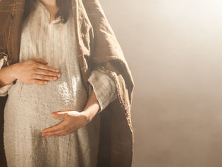 A person in a brown shawl and textured white dress cradles their pregnant belly. Soft warm lighting creates a peaceful mood.