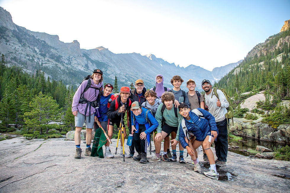 Group of hikers posing on a rocky mountain landscape with green forests and peaks. They are smiling under a clear blue sky.
